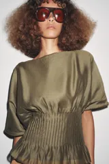 Blusa de manga corta con escote barco, confeccionada en tejido satinado color verde oliva. Presenta un diseño holgado en la parte superior y un canesú inferior con pliegues verticales ajustados.