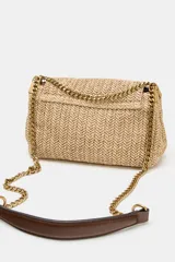 Bolso de hombro formato bandolera con cuerpo en efecto trenzado de color beige. Cuenta con asa de hombro combinada de cadena metálica dorada y tira de cuero sintético marrón, compartimento interior con cremallera y cierre mediante botón imantado.