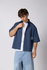 Camisa de manga corta negra 100% algodón, con calce boxy, cuello camisero, cierre frontal con botones y detalle de bordado blanco en la parte inferior.