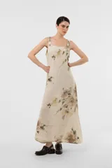 Vestido largo de lino color beige, con estampado floral en tonos marrones y verdes. Cuenta con escote redondo, tirantes anchos con detalle de botones decorativos y una silueta fluida y amplia.