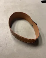 Cinturón ancho de cuero negro con doble hilera de tachas plateadas. Hebilla metálica plateada de estilo occidental y punta de cierre en forma de flecha.