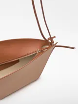 Bolso de hombro Adolfo Dominguez modelo Eclipse, confeccionado en piel bovina color tostado. Presenta un diseño estructurado formado por dos piezas asimétricas entrecruzadas, cierre con cremallera, asa de hombro extraíble y logo grabado en la parte frontal.