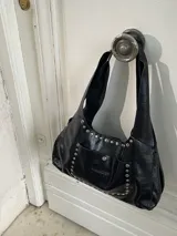 Bolso tipo tote de cuero color visón con apliques de tachas plateadas. Tiene doble asa corta, cierre con imán y bolsillo externo con imán. Incluye un sobre interno grande con gancho.