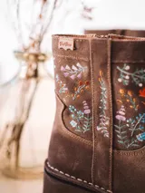 Botín de mujer Monells elaborado en cuero serraje color beige, con tacón ancho y detalles de flores bordadas en el lateral. Cuenta con cierre de cremallera lateral.