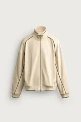 Chaqueta estilo deportivo de corte relajado, color beige con detalles de vivos en color verde oscuro. Presenta cuello alto, cierre frontal con cremallera de doble dirección, bolsillos laterales y puños elásticos.