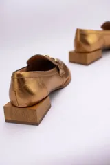 Mocasines de cuero en tono bronce metalizado, con detalle de aplique metálico dorado en el empeine y taco cuadrado bajo de madera.