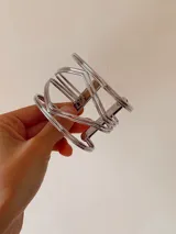 Brazalete abierto de diseño geométrico en tono plateado, compuesto por múltiples líneas metálicas que forman una estructura central en forma de rombo.