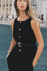 Chaleco negro de vestir, sin mangas, con cuello redondo y cierre frontal de botones metálicos dorados. Incluye dos bolsillos frontales con solapa y un cinturón negro a tono con hebilla metálica dorada.