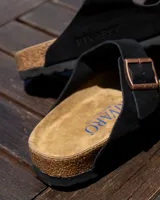 Sandalias tipo zueco marca Rivaro, confeccionadas en cuero gamuzado negro con dos tiras ajustables con hebillas metálicas. Presentan una plantilla anatómica de corcho y látex, y suela de goma antideslizante.