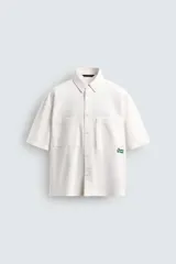Camisa de algodón color blanco crudo, con cuello solapa, manga corta y dos bolsillos de plastrón en el pecho. Presenta un bordado y estampado de texto en color verde en el lado derecho del pecho.