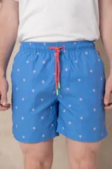 Short de baño azul vibrante con estampado repetitivo de cruces pequeñas en blanco y rojo. Presenta cintura elástica fruncida y cordón de ajuste trenzado en color coral con puntas de neón verde lima.
