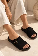 Sandalias planas de tipo chinela en color negro, confeccionadas con una tira ancha sobre el empeine que incluye una hebilla metálica dorada decorativa y una abertura en la puntera.