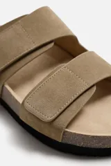 Sandalia color beige fabricada en piel con acabado serraje. Cuenta con dos tiras anchas ajustables mediante cierres adherentes y plantilla con horma anatómica.