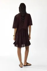 Vestido corto de color bordó, con escote en V y cuello de volados, mangas cortas abullonadas y botonadura frontal con alforzas e inserciones de encaje. Presenta costuras angulares con frunces en la cadera y falda con ribete de volado en el bajo.