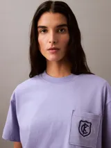 Remera lila de corte cropped y boxy, con cuello redondo y bolsillo en el pecho con escudo bordado en negro.
