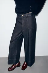 Pantalón de jean tiro medio con corte culotte, pernera ancha y corta. Presenta bolsillos delanteros de plastrón, costuras decorativas verticales y cierre frontal con cremallera y botón.