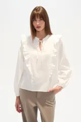 Blusa blanca de algodón con volados frontales, cuello redondo con lazo ajustable y mangas largas abullonadas con puños elásticos.
