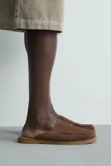 Zueco de piel con acabado serraje en color marrón, diseño sin talón, puntera redonda y detalle de costura tipo mocasín en el empeine.