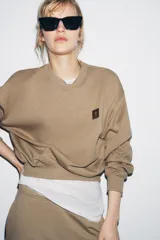 Sudadera de algodón color beige, con cuello redondo, manga larga y corte ligeramente corto. Presenta un pequeño parche cuadrado marrón con un logo grabado en el pecho.