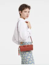 Cartera tipo crossbody de cuero color terracota, con forma cilíndrica, solapa frontal con cierre de botón a presión metálico y correa ajustable. Incluye un colgante decorativo de cuentas coloridas en uno de los laterales.