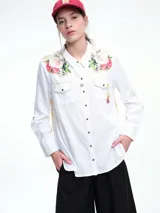 Camisa blanca de corte clásico con bordados florales en los hombros y detalles decorativos en los bolsillos frontales. Cuenta con cierre de botones y mangas largas.