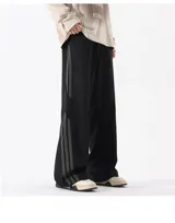 Pantalón deportivo negro de corte ancho y tiro alto con cordón ajustable en la cintura. Presenta tres franjas verticales grises oscuras en los laterales y un bolsillo cargo con solapa en el muslo derecho. Los tobillos son ajustables.