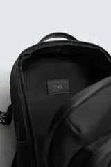 Mochila técnica de color negro con múltiples compartimentos, incluyendo dos bolsillos frontales con cierre, bolsillos laterales y compartimento principal acolchado para dispositivos electrónicos. Cuenta con parte posterior acolchada, banda de sujeción para equipaje, asa de mano y asas de hombro ajustables.
