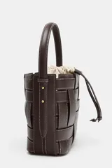 Bolso tipo bucket de diseño trenzado en color marrón oscuro con costuras a la vista en contraste. Cuenta con asa de mano fija, correa bandolera extraíble y compartimento interior de tela en color crudo con cierre mediante fruncido.