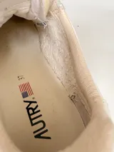 Championes Autry de diseño retro, color blanco con detalles en gamuza beige y suela de tono tostado, con cordones claros y logo lateral de la marca con la bandera de Estados Unidos.