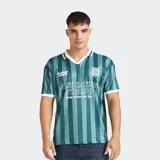 Remera estilo camiseta de fútbol con cuello polo, manga corta y diseño de rayas verticales en tonos verdes. Presenta estampados gráficos en el frente y escudo aplicado en el pecho.