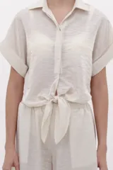 Camisa crop fluida de manga corta color crema con cuello camisero, botonadura frontal oculta y detalle de nudo en el frente.
