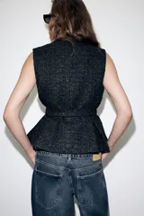 Chaleco de tejido tipo tweed en color negro, con cuello redondo y cierre frontal de botones dorados. Presenta un diseño entallado con cinturón integrado del mismo tejido y bajo con pliegues.