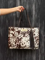Cartera tipo tote de cuero con estampado animal print tipo vaca en tonos marrón y blanco. Cuenta con doble asa de hombro en cuero marrón oscuro, cierre de broche metálico dorado y un detalle de borla colgante de flecos de cuero.