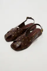 Sandalias planas de cuero sintético marrón, con tiras cruzadas en el empeine y tachas metálicas doradas distribuidas por toda la capellada. Cuenta con tira ajustable en el talón con hebilla.