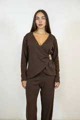 Blusa de color marrón con acabado brillante, diseño cruzado tipo sobre con escote en V y manga larga. Presenta un detalle de broche decorativo en forma de sol en el lateral de la cintura.