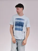 Remera celeste de manga corta y cuello redondo, con estampado fotográfico frontal de un mar y el texto "We are moved by the sea".
