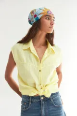 Camisa de lino color amarillo manteca, sin mangas, con cuello clásico y cierre frontal con botones. Presenta un diseño corto con nudo en el ruedo.