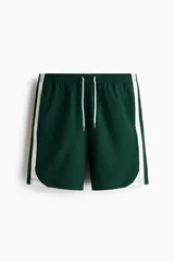 Short deportivo de color verde oscuro, confeccionado en nylon, con bandas laterales blancas, cintura elástica ajustable mediante cordón blanco y corte estándar.