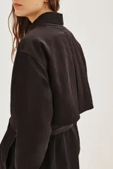 Trench negro de corte midi con cuello camisero, cierre frontal y cinturón ajustable a la cintura. Presenta mangas largas con puños rectos y un diseño minimalista y versátil.