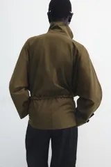 Parka amplia de Zara Woman Collection confeccionada en hilatura con mezcla de algodón, en color verde militar. Presenta cuello alto, manga larga con trabillas y botón, bolsillos laterales ocultos en costura, cintura ajustable con cordones y cierre frontal con botones.
