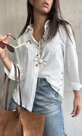 Camisa de lino color blanco, de corte holgado y manga larga con puños arremangados. Presenta cuello clásico, cierre frontal con botones y detalle de botones decorativos en el lateral inferior.