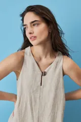 Vestido corto de lino color beige, de silueta suelta y sin mangas, con escote en V decorado con un botón, bolsillos laterales y detalle de volado en el ruedo.