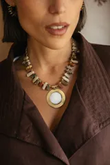 Collar corto con cadena compuesta por bastones de piedras naturales en tonos tierra y mostacillas, con un medallón central circular de nácar engarzado en metal dorado.