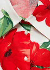 Camisa de corte crop con cuello camisero y cierre frontal de botones, destacada por su diseño estampado de flores rojas sobre fondo blanco. Presenta mangas largas con puños abotonados y un detalle de lazo anudado en la parte inferior delantera.