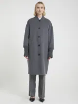 Abrigo largo de color gris confeccionado a mano, con cuello y puños acanalados. Presenta un diseño de corte recto con cierre frontal de botones, bolsillos laterales y botones decorativos en la parte posterior.