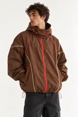 Campera rompevientos oversize color marrón, de nylon, con cuello alto, cierre frontal asimétrico con cremallera roja y detalles de ribetes en contraste.