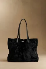 Bolso tipo tote de gran tamaño en color negro, confeccionado en material con textura similar a la gamuza. Presenta doble asa larga para llevar al hombro y una placa metálica con el logo de la marca en la parte frontal.