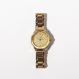 Reloj de pulsera dorado con malla metálica, caja octogonal y esfera dorada con agujas y marcadores de hora minimalistas.