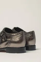 Zapato tipo monk strap con aberturas laterales, confeccionado en cuero vacuno metalizado color bronce. Presenta detalles de perforaciones estilo brogue en la puntera y el talón, y doble hebilla metálica en el empeine.