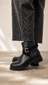 Bota corta de cuero negro con suela track de goma, detalle de tira lateral con hebilla metálica y acabado minimalista.